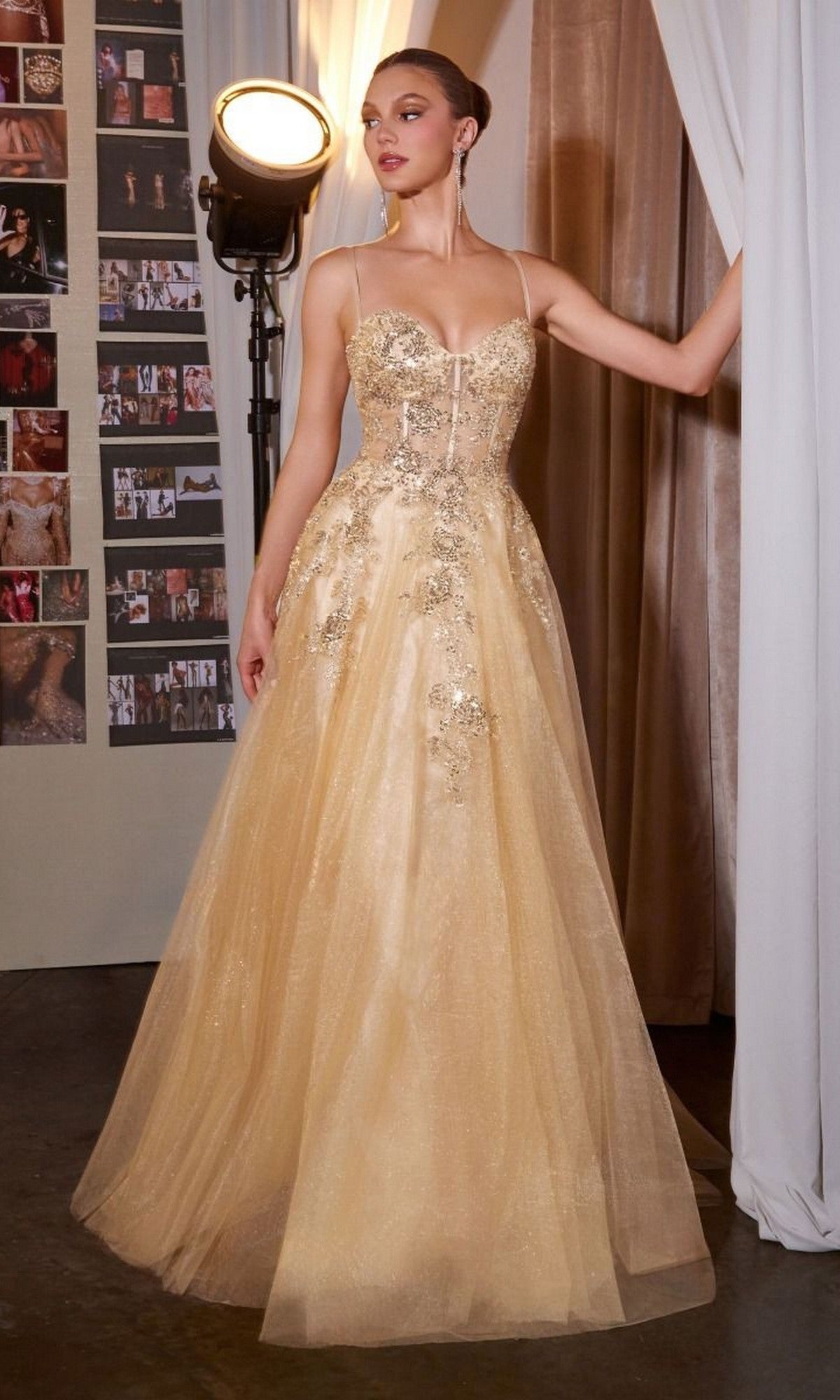 A woman in the Ladivine C177 long prom dress in gold, featuring an embellished bodice and thin straps, stands indoors near a curtain and spotlight with photo collages on the wall behind her.